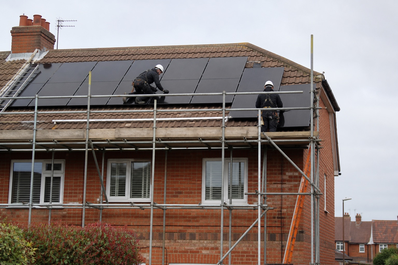 Solar installers fitting black panels to a tiled roof on a red-brick UK house using scaffolding